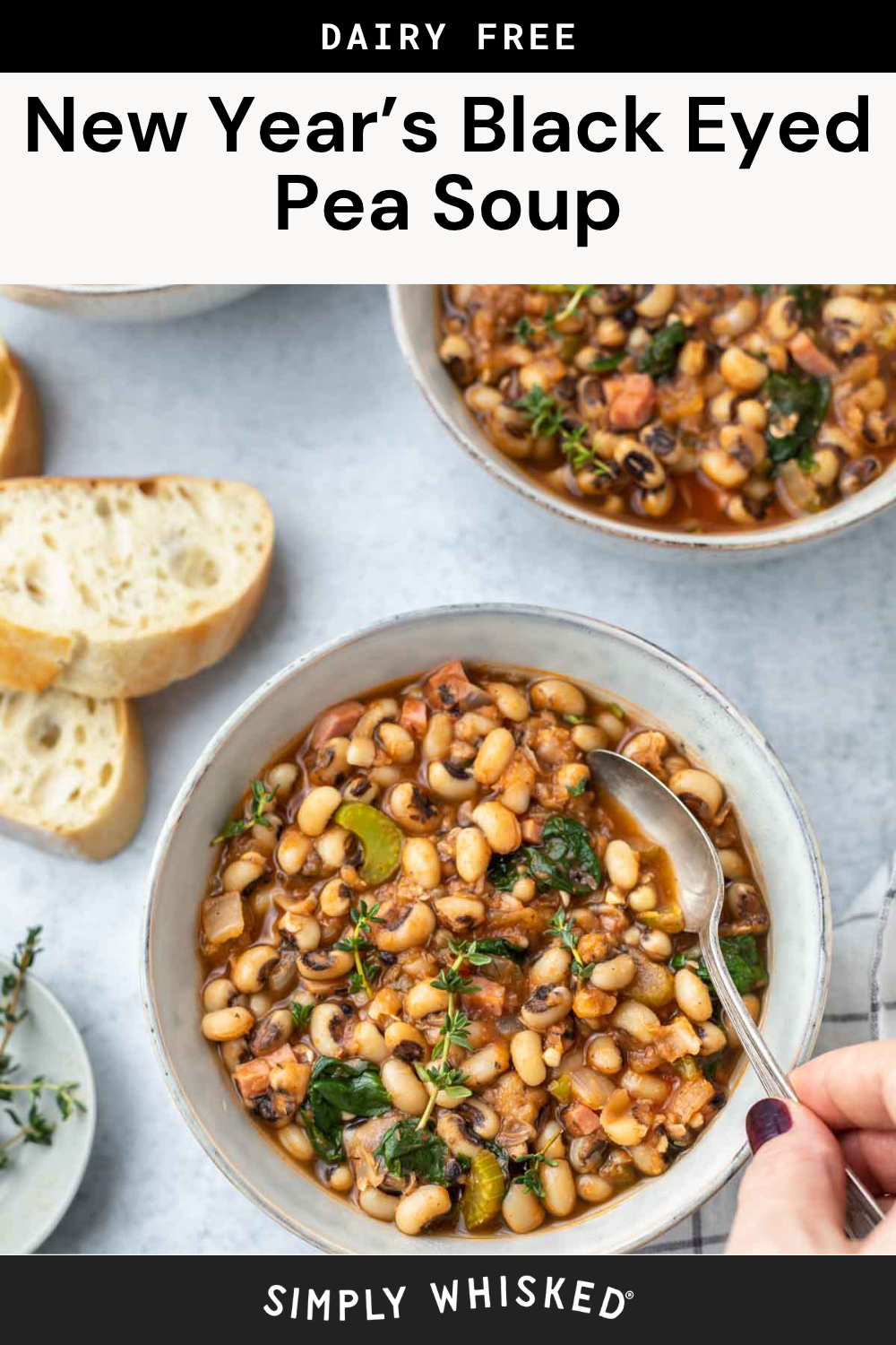 A steaming bowl of black eyed pea soup with greens and vegetables sits on a table next to slices of bread, fresh herbs, and a spoon, with another bowl visible in the background.