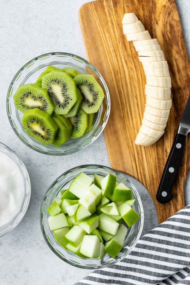 a bowl of chopped apple and a bowl of sliced kiwi next to a cutting board with a sliced banana
