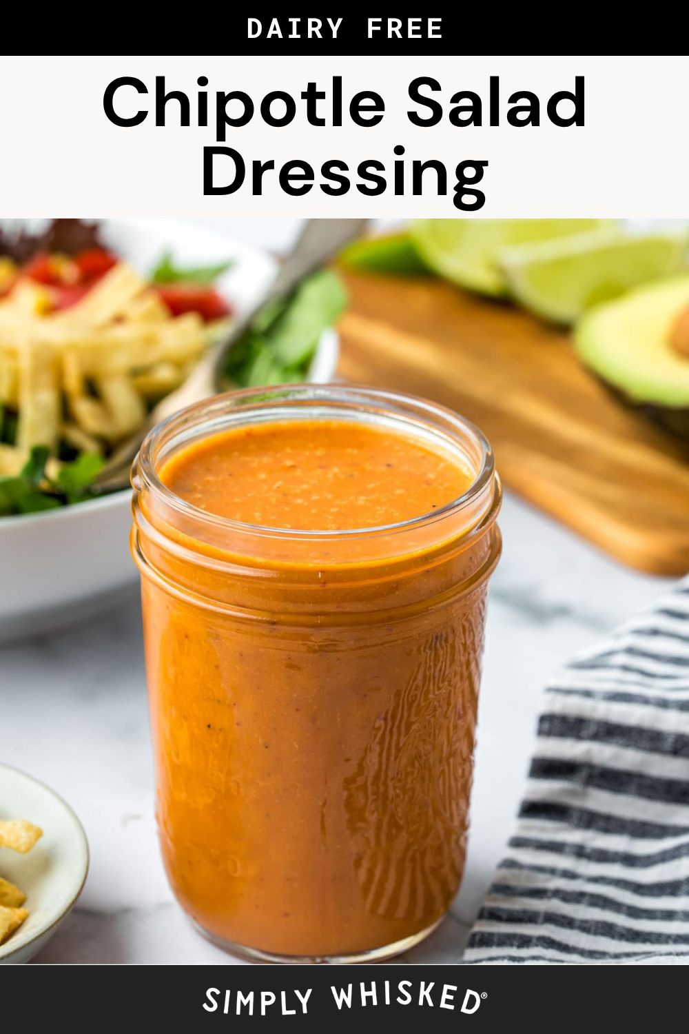 A glass jar filled with vibrant orange chipotle dressing sits on a white surface, accompanied by a striped towel, fresh salad, and sliced avocado in the background. Text above the jar reads Dairy Free Chipotle Salad Dressing.
