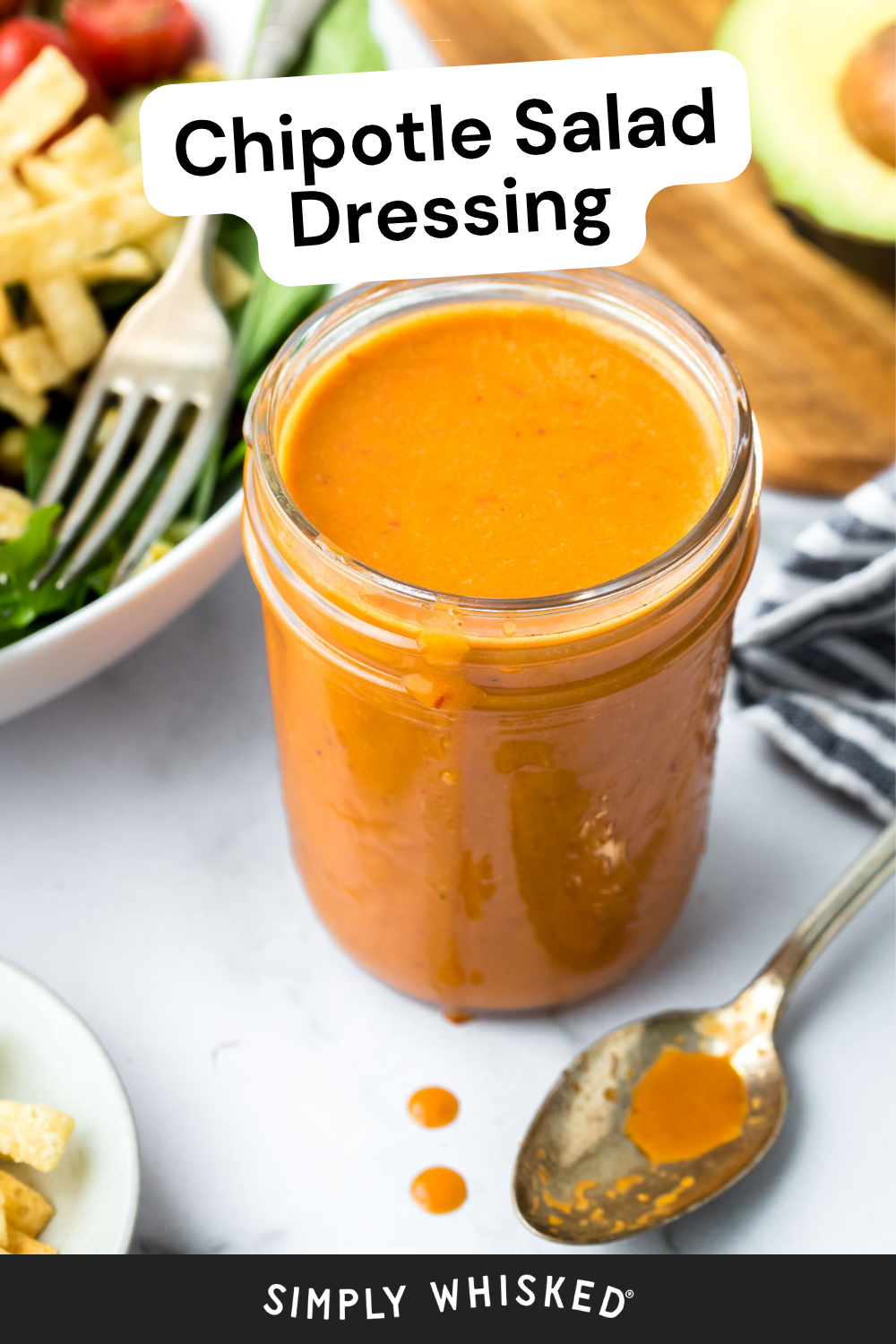 A jar filled with vibrant orange chipotle dressing sits on a white surface, with a spoon holding some of the smoky salad dressing nearby. Part of a fresh salad and half an avocado are visible in the background.