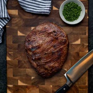 A whole marinated flank steak on a cutting board with a knife, towel, and garnish off to the side