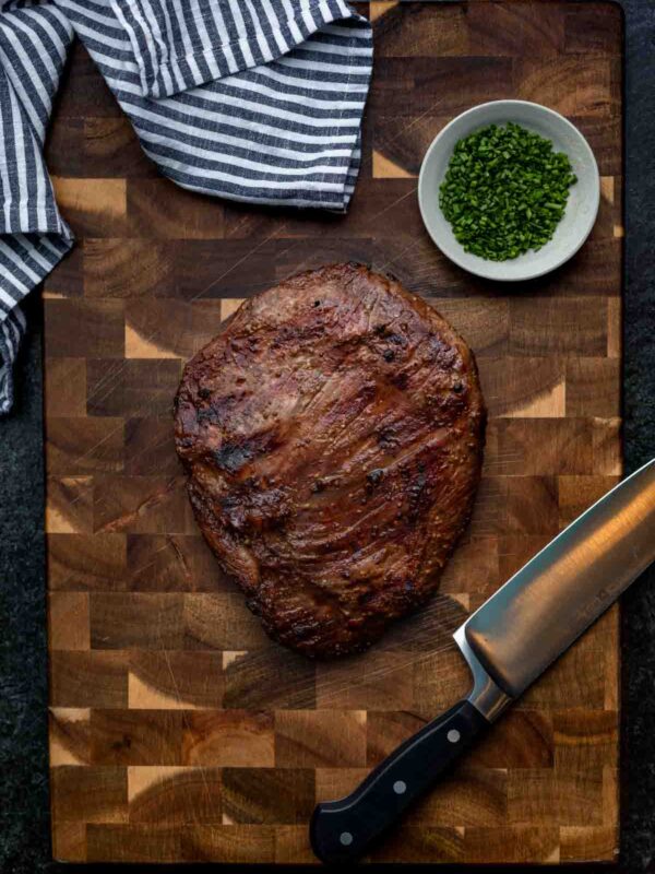 A whole marinated flank steak on a cutting board with a knife, towel, and garnish off to the side