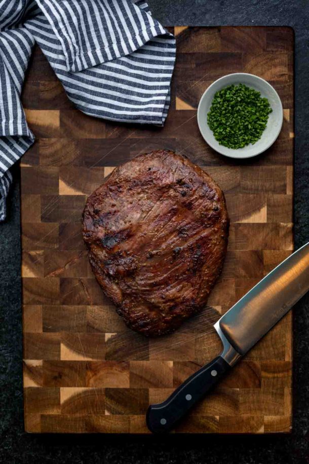 A whole marinated flank steak on a cutting board with a knife, towel, and garnish off to the side