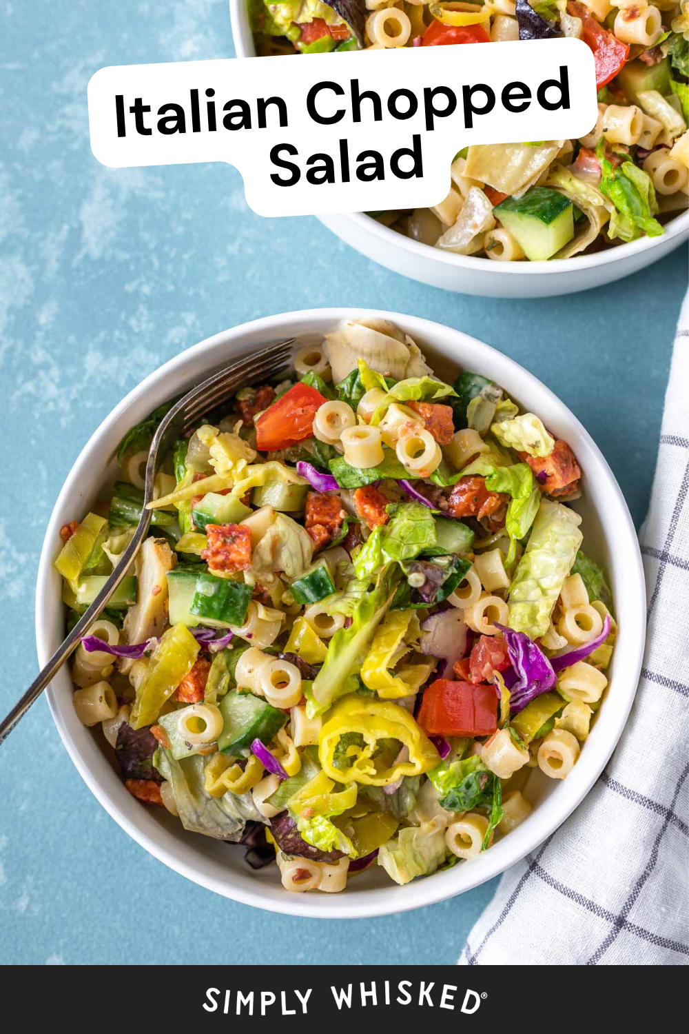 A bowl of Italian chopped salad with pasta, lettuce, tomatoes, cucumbers, and pepperoncini sits invitingly on a blue surface alongside a fork. A partial second bowl of Italian chopped salad and a striped napkin are also visible in the scene.
