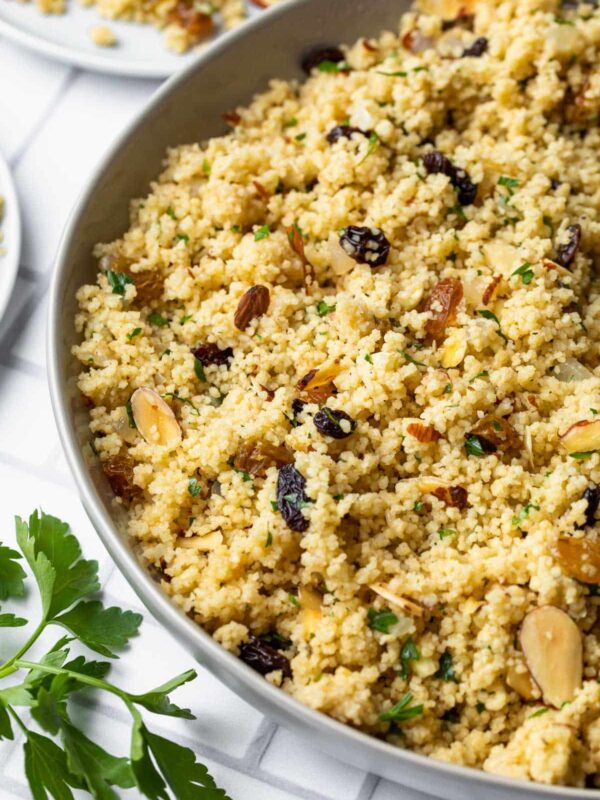 A bowl of Moroccan couscous mixed with sliced almonds, raisins, and chopped herbs sits on a white checkered surface, with a sprig of parsley nearby.