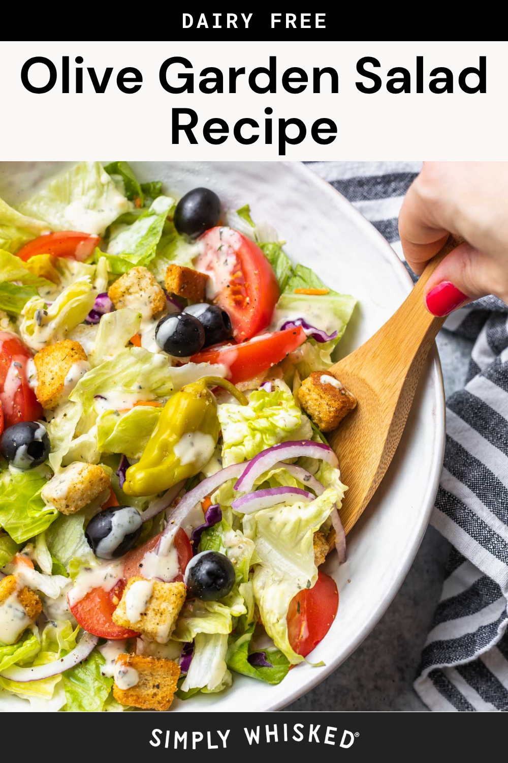 A bowl of salad with lettuce, tomatoes, black olives, croutons, peppers, and red onions—just like an Olive Garden salad. A hand uses a wooden spoon to serve, and creamy dressing is drizzled on top.