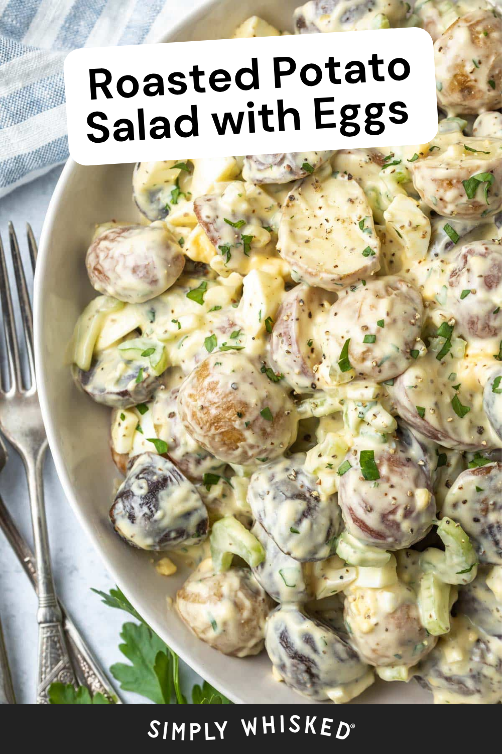 A bowl of roasted potato salad with eggs, celery, and herbs in a creamy dressing, garnished with parsley. Two forks are placed next to the bowl on a striped towel.