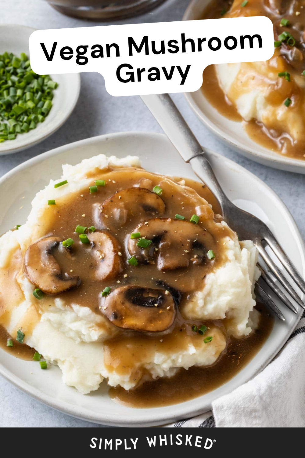 Plate of mashed potatoes topped with rich mushroom gravy and chopped chives, with a fork and knife on the side. Another plate and a bowl of chopped chives are in the background.