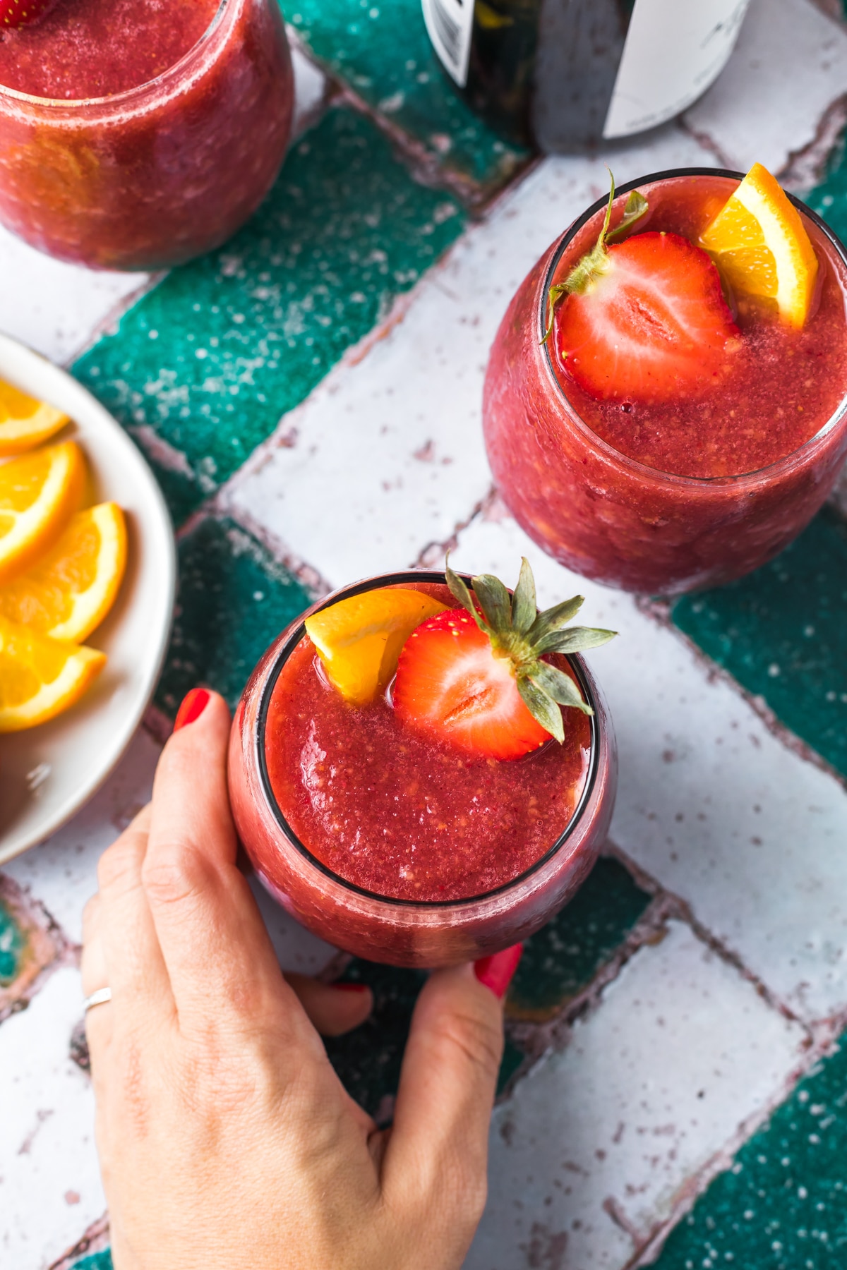 over head view of hand reaching for glass of frozen sangria with orange and strawberry slices, small plate with orange slices and second glass in view