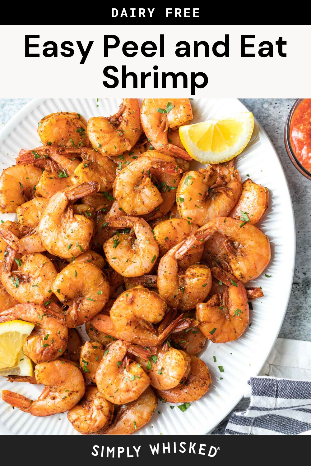 A white plate of seasoned, cooked peel and eat shrimp is garnished with fresh herbs and lemon wedges, set beside classic cocktail sauce. The text above reads “Easy Peel and Eat Shrimp” and “DAIRY FREE.”.