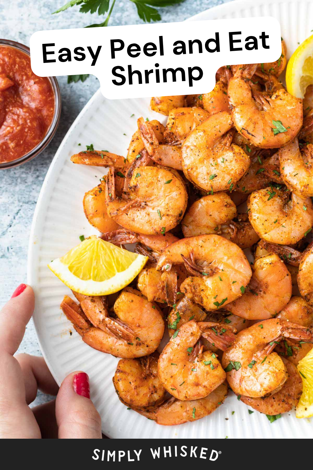 A plate of seasoned peel and eat shrimp garnished with lemon wedges, with a hand reaching in and cocktail sauce nearby. Text reads Easy Peel and Eat Shrimp and Simply Whisked.
