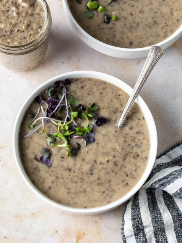 A bowl of vegan cream of mushroom soup topped with microgreens, with a spoon in the bowl. Nearby, another bowl, a jar of soup, and a striped cloth napkin rest on a light surface.