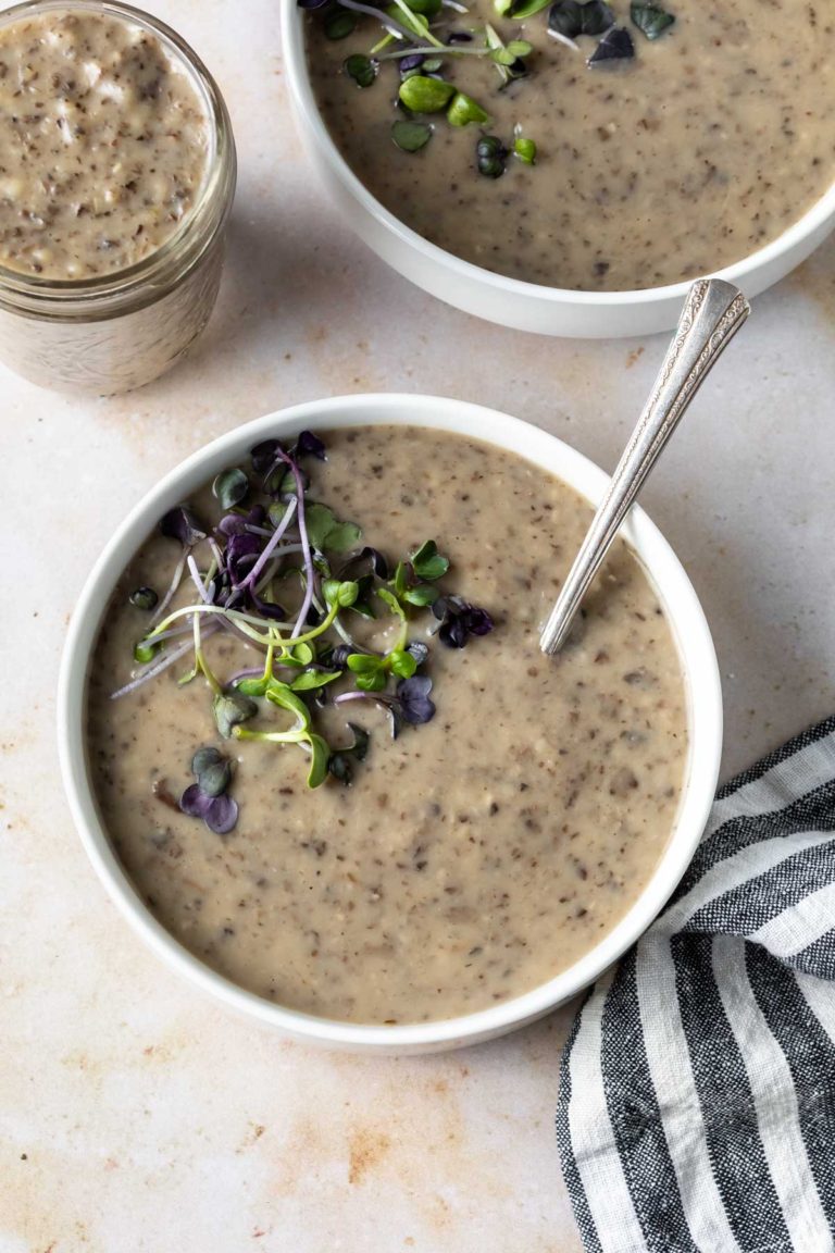 A bowl of vegan cream of mushroom soup topped with microgreens, with a spoon in the bowl. Nearby, another bowl, a jar of soup, and a striped cloth napkin rest on a light surface.