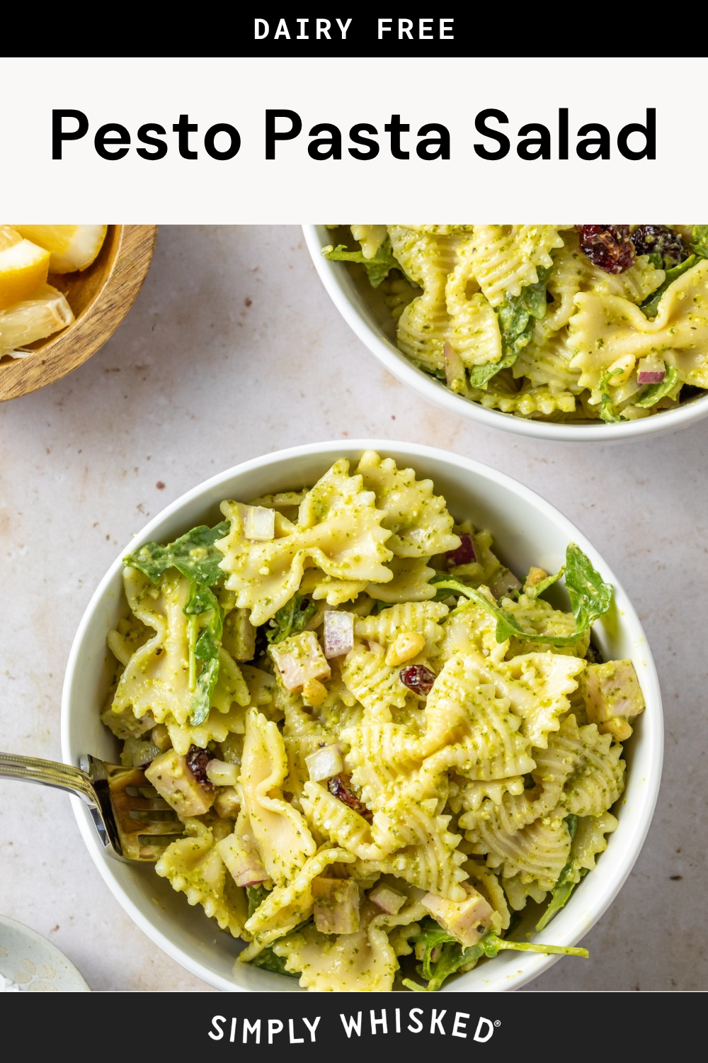 Two white bowls filled with vibrant pesto pasta salad, featuring bowtie pasta tossed with greens, red onion, and sun-dried tomatoes. A fork rests in one bowl, while a wooden bowl of lemon wedges sits nearby.