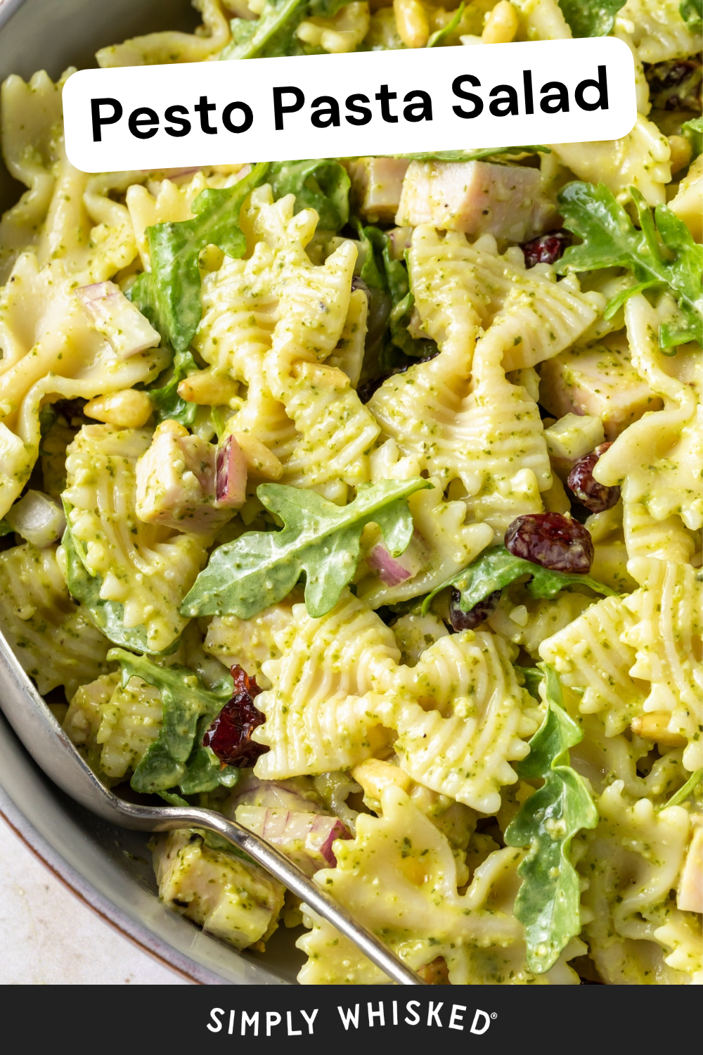 A close-up of pesto pasta salad with bowtie pasta, arugula, cranberries, red onion, and chicken cubes. A serving spoon is in the bowl. Text overlay reads Pesto Pasta Salad and Simply Whisked. This vibrant pesto pasta salad makes for a fresh and flavorful meal or side dish.