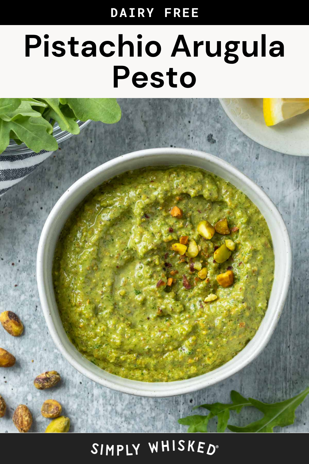 A bowl of vibrant pistachio arugula pesto is topped with chopped pistachios and surrounded by whole pistachios, fresh arugula leaves, and a lemon wedge on a gray surface.
