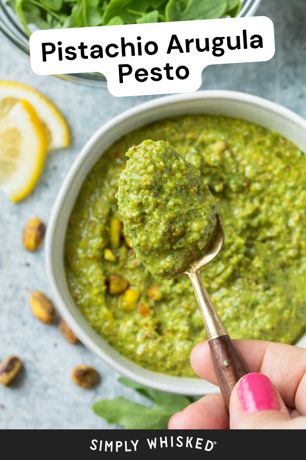 A hand holds a spoonful of vibrant pistachio arugula pesto above a bowl, with scattered pistachios, fresh arugula, and lemon slices creating a colorful background.