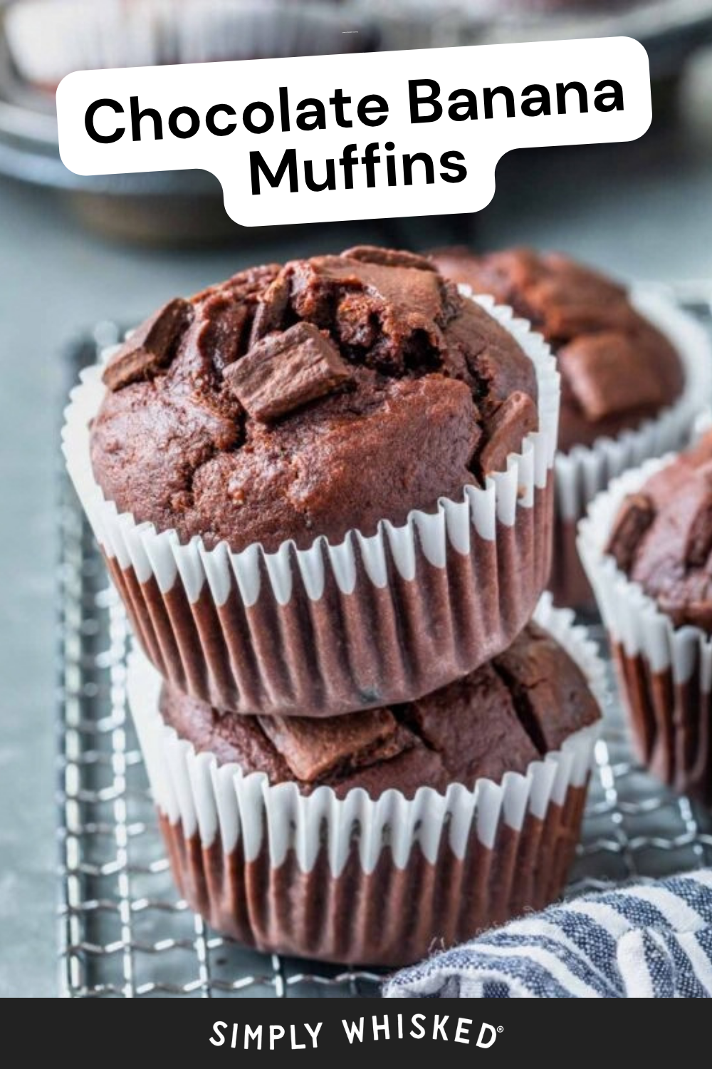 Two stacked chocolate banana muffins with chocolate chunks, in white wrappers, on a cooling rack. More muffins and a striped cloth are visible in the background. Text on the image reads, Chocolate Banana Muffins..