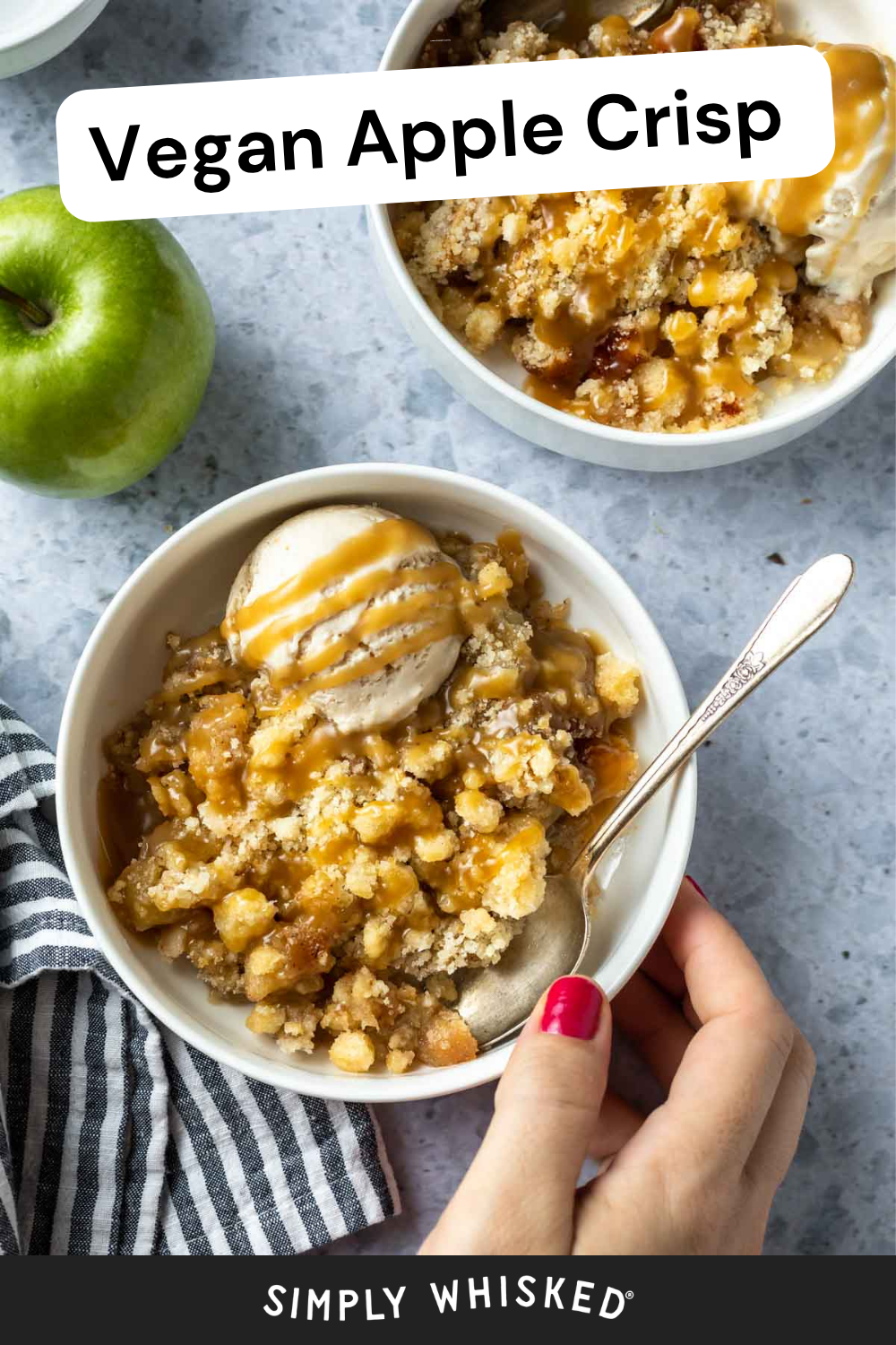 A bowl of vegan apple crisp topped with vanilla ice cream and caramel sauce, with a spoon and a hand holding the bowl. Another bowl of vegan apple crisp, a green apple, and a striped towel are nearby.