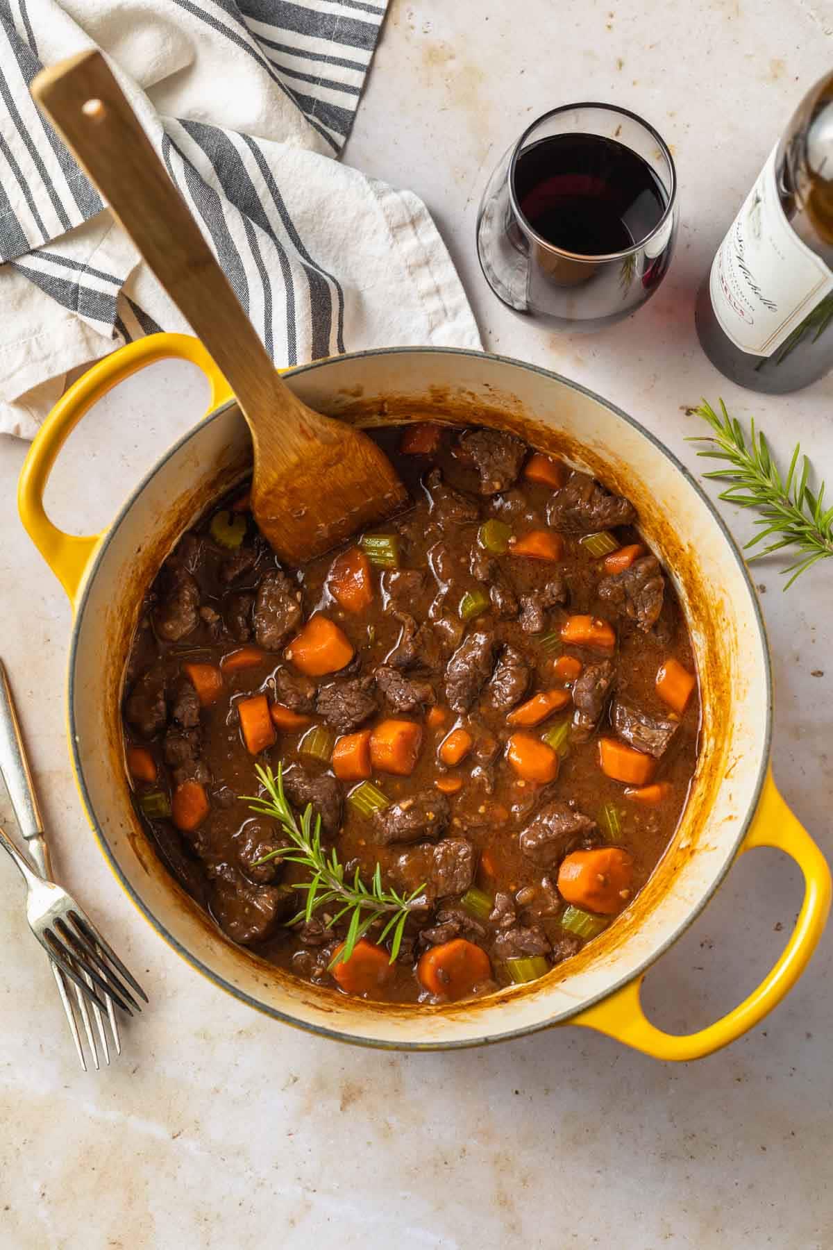 overhead view of wooden spoon staring a dutch oven full of red wine beef stew on a countertop next to a glass of red wine