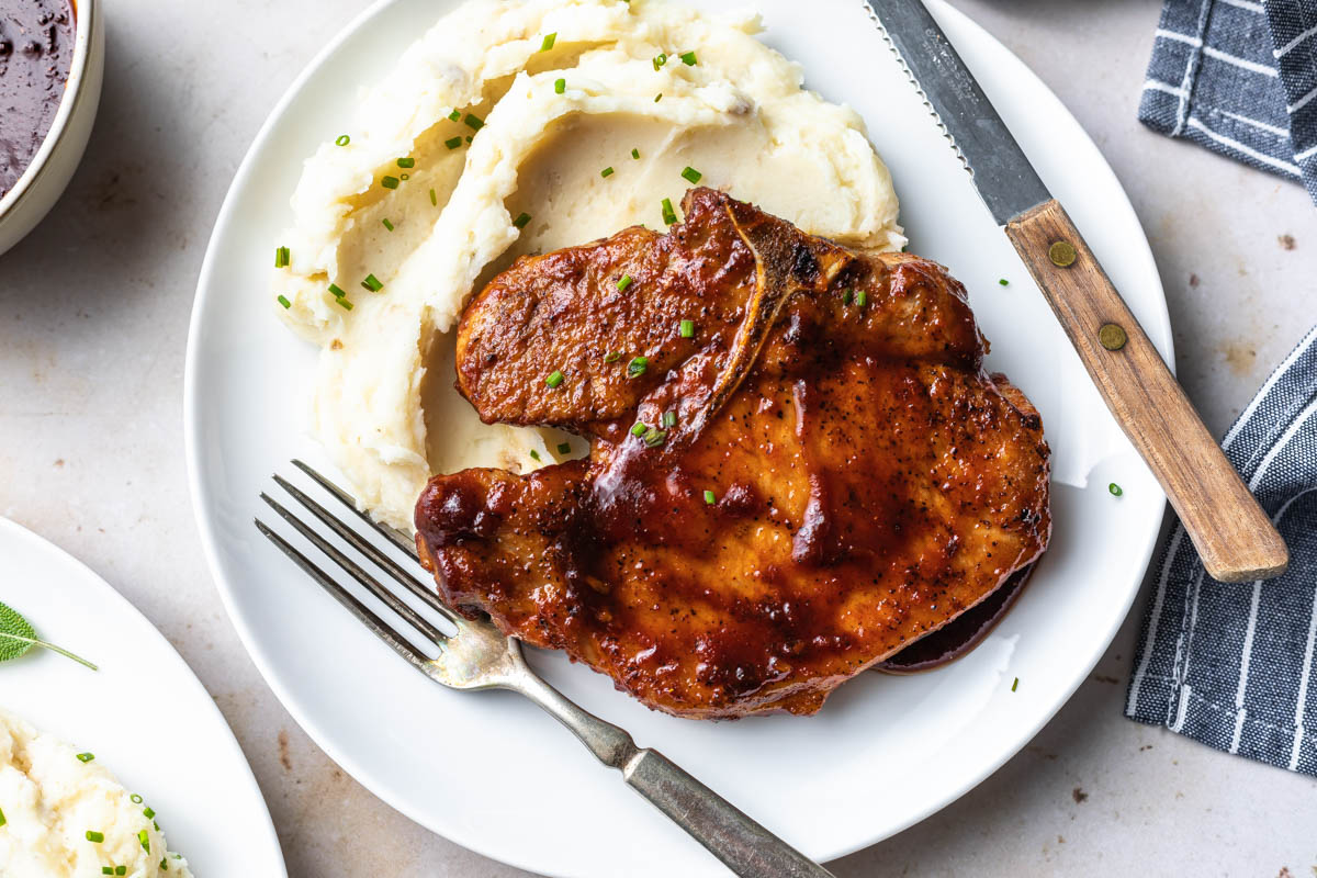 glazed pork chop on a bed of mashed potatoes with a fork and knife on a plate