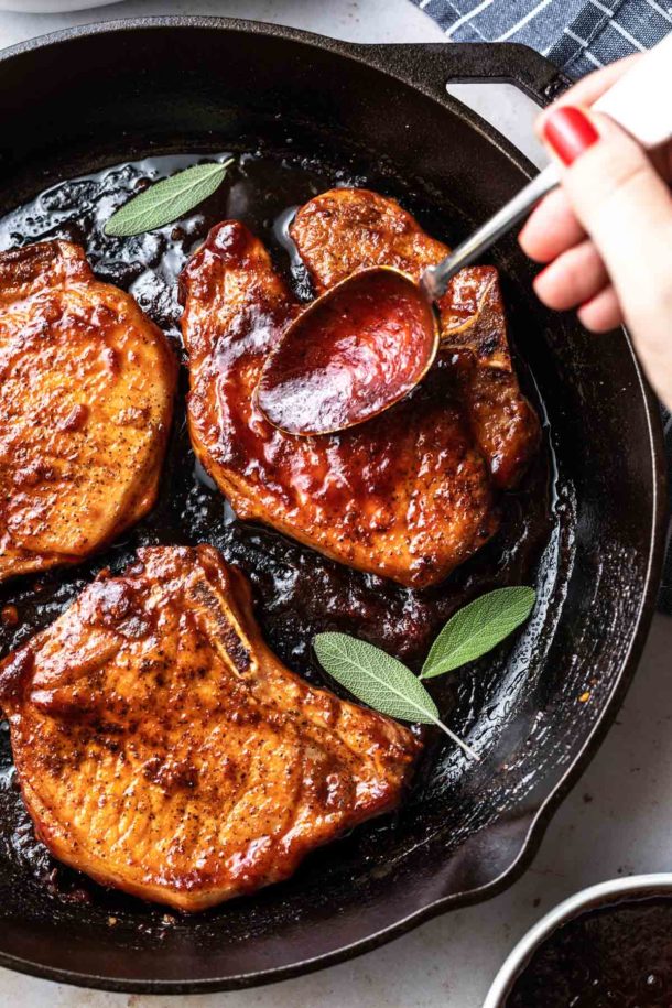 A hand spoons barbecue sauce over three glazed pork chops in a cast iron skillet, garnished with fresh sage leaves.