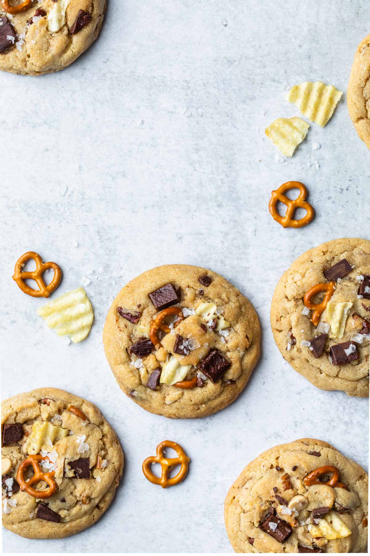 Kitchen sink cookies packed with chocolate chips, pretzel twists, and ridged potato chips are arranged on a light gray surface, with extra pretzels and chips scattered around the cookies.