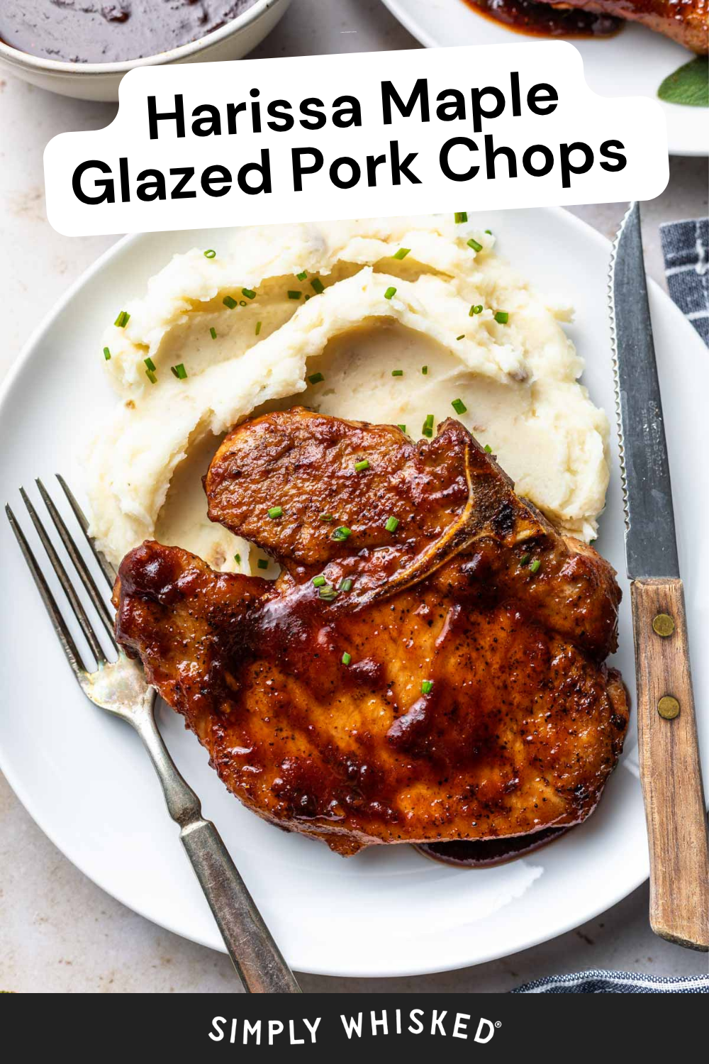 A plate with two glazed pork chops, mashed potatoes topped with chopped chives, and a knife and fork resting on the plate. The dish is labeled Harissa Maple Glazed Pork Chops, showcasing perfectly glazed pork chops paired with creamy, flavorful sides.