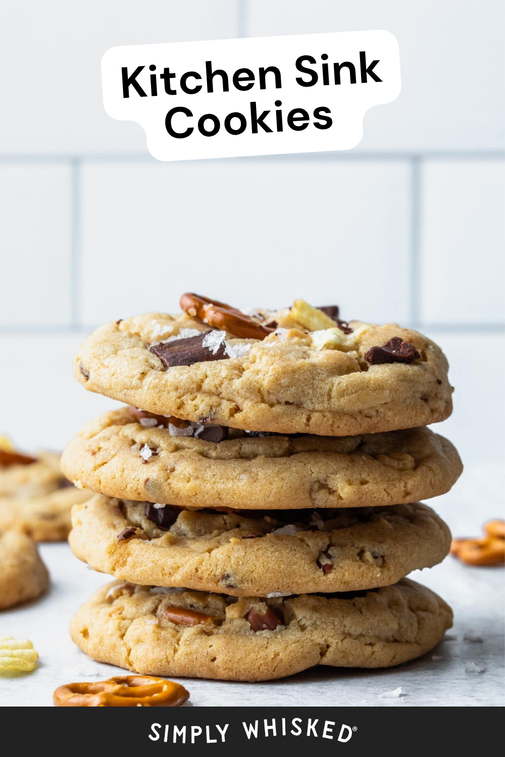 A stack of four thick kitchen sink cookies, loaded with chocolate chunks and nuts, is labeled Kitchen Sink Cookies. The cookies rest on a white surface with pretzels and nuts scattered nearby. Text at the bottom reads Simply Whisked.