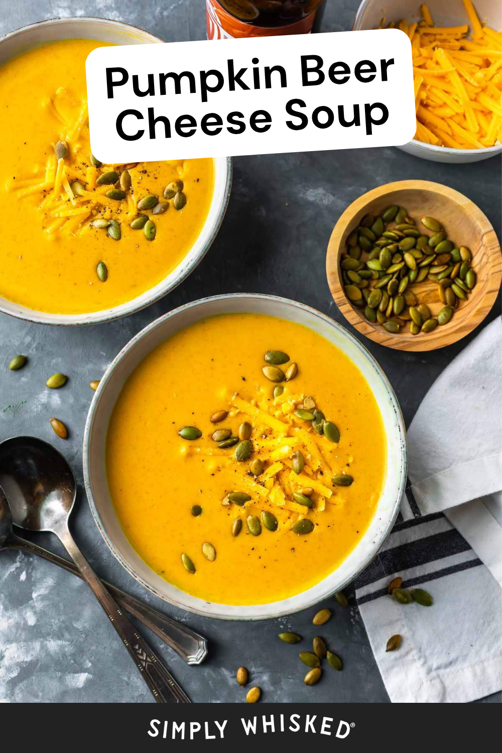 Two bowls of beer cheese soup topped with shredded cheese and pumpkin seeds, next to a bowl of seeds, a spoon, and a striped napkin. Text overlay reads Pumpkin Beer Cheese Soup.
