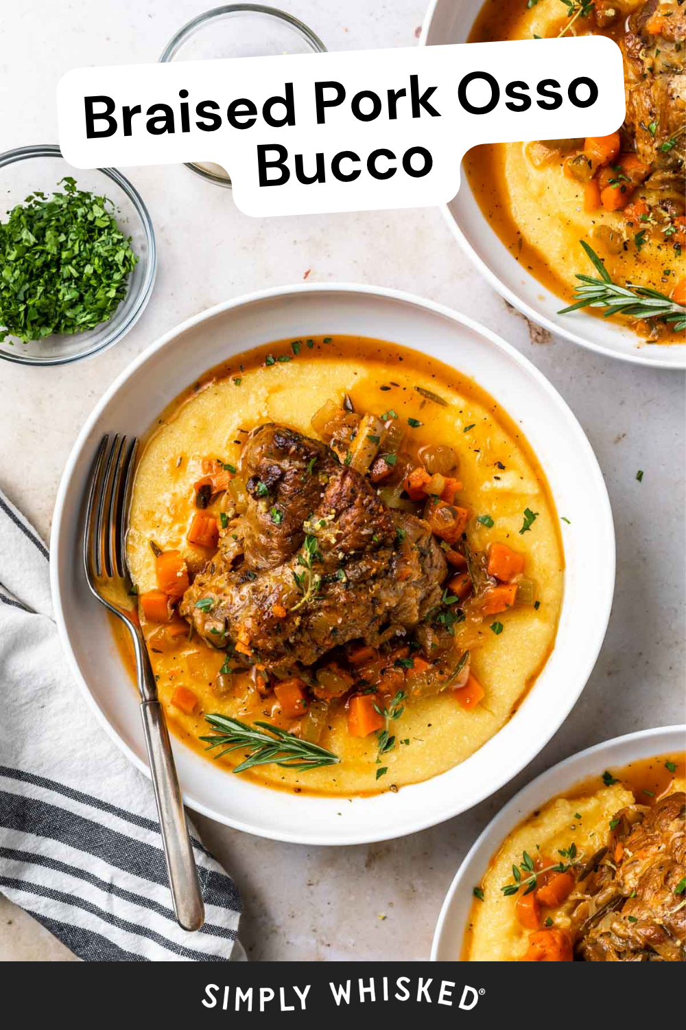 A bowl of tender pork osso bucco is served over creamy polenta, topped with carrots and herbs. A fork and a small bowl of chopped parsley are beside the plate on a striped napkin.