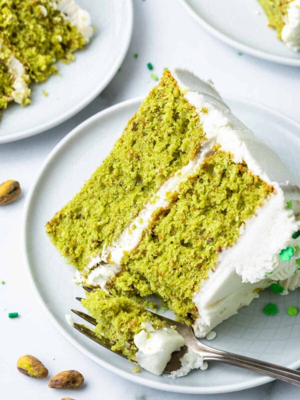 A slice of pistachio cake with green layers and white frosting sits on a white plate, a forkful missing. Whole pistachios and other pistachio cake slices are scattered nearby on a clean white surface.