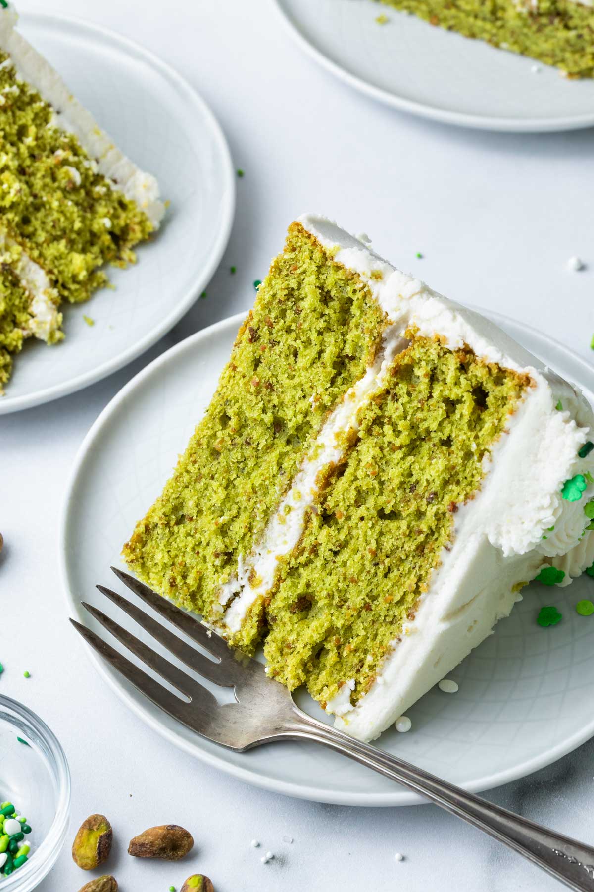 A slice of green nut flavored cake with white frosting, decorated with green sprinkles. 