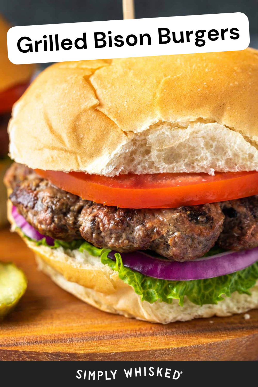 Close-up of a juicy grilled bison burger on a bun with fresh lettuce, red onion, and tomato. A pickle is on the side. Text at the top reads Grilled Bison Burgers—perfect for anyone craving bold and flavorful bison burgers.
