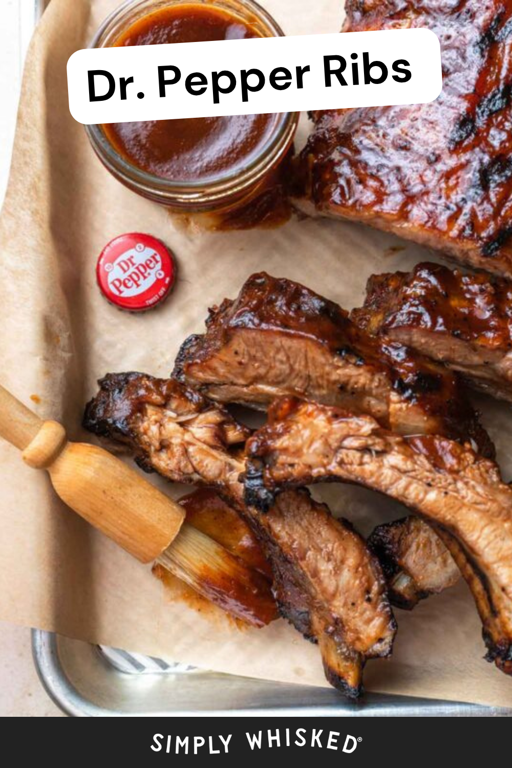 Ribs glazed with sauce on parchment, a basting brush, a small jar of sauce, and a Dr. Pepper bottle cap set the scene for these mouthwatering Dr. Pepper ribs. Text reads Dr. Pepper Ribs. Bottom text: Simply Whisked.