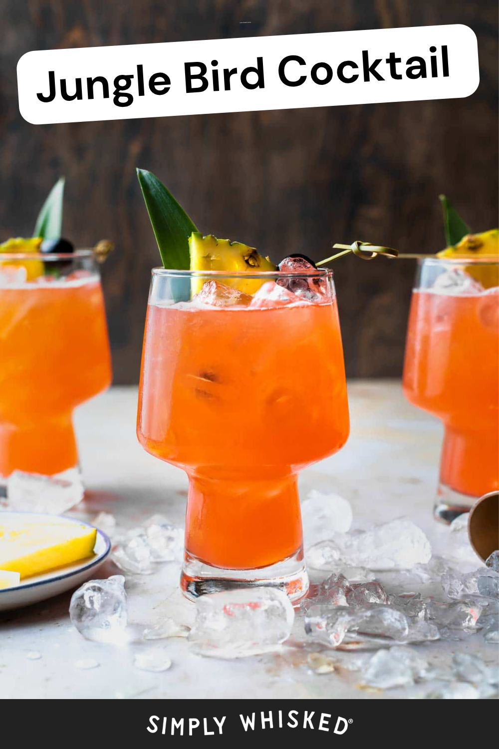 Three orange Jungle Bird Cocktails with ice, garnished with pineapple, cherry, and greenery, sit on a table scattered with ice cubes. The vibrant scene features "Jungle Bird Cocktail" text above and "Simply Whisked" below.