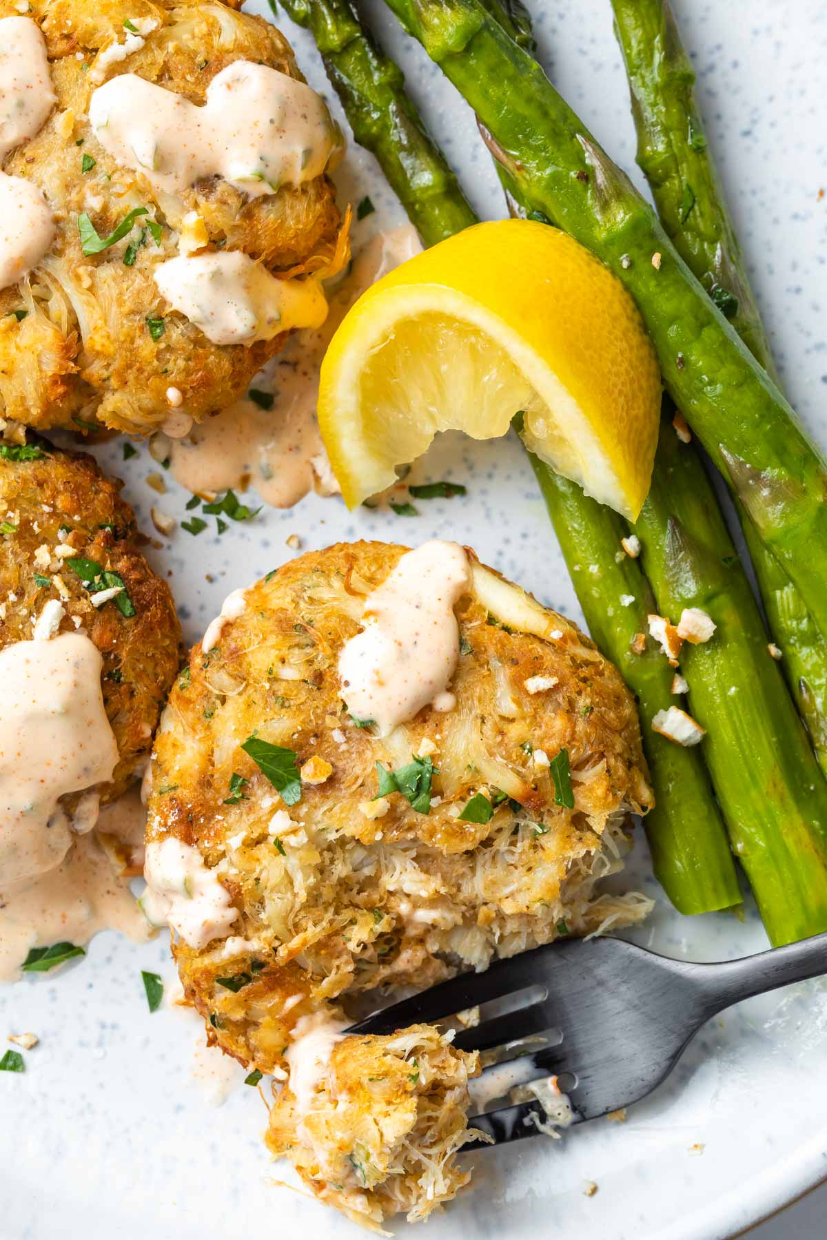 Close up of 3 crab cakes drizzles with a cajun remoulade on a plate with asparagus.