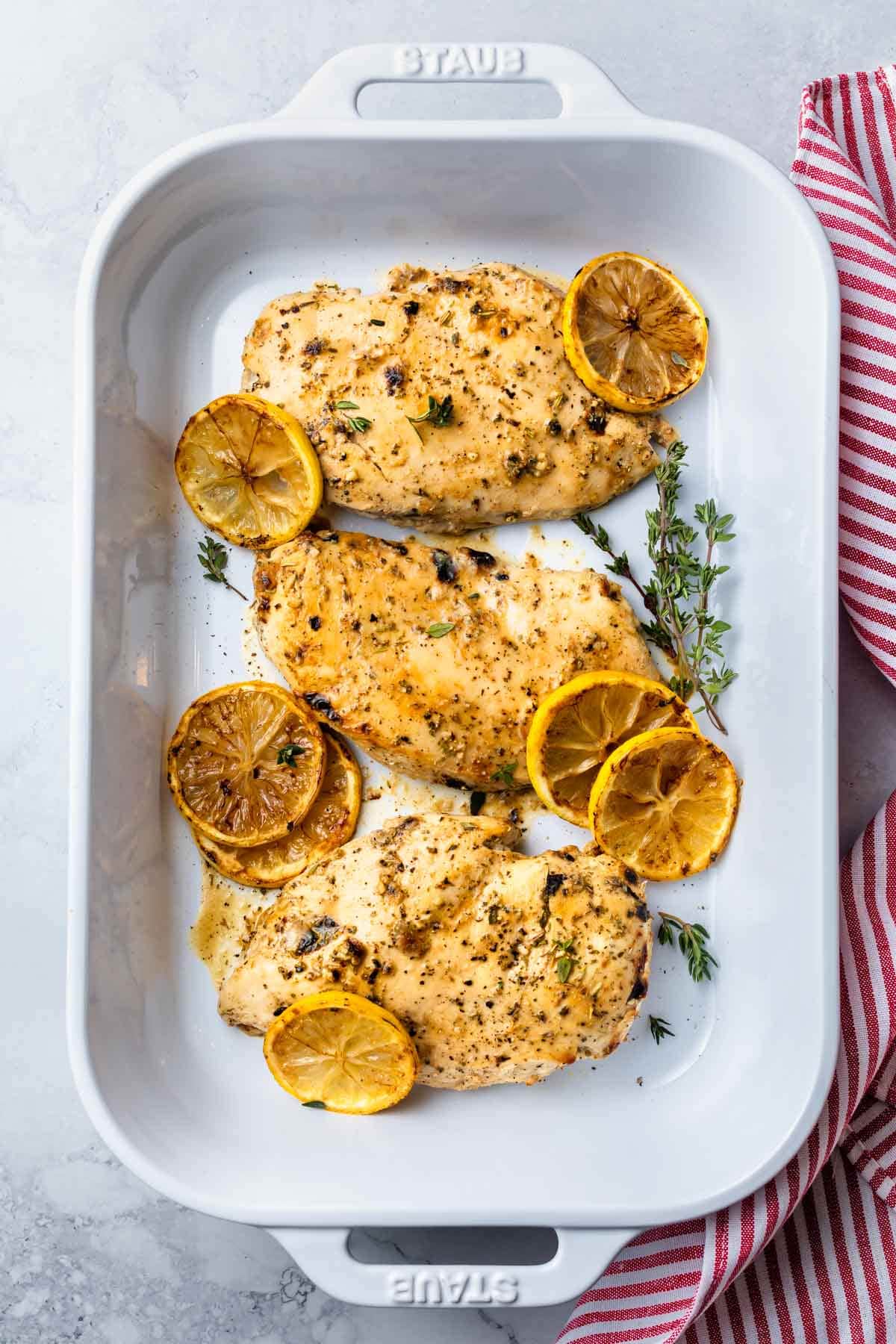 Three chicken breast in a white baking dish with sliced lemons and springs of herbs.