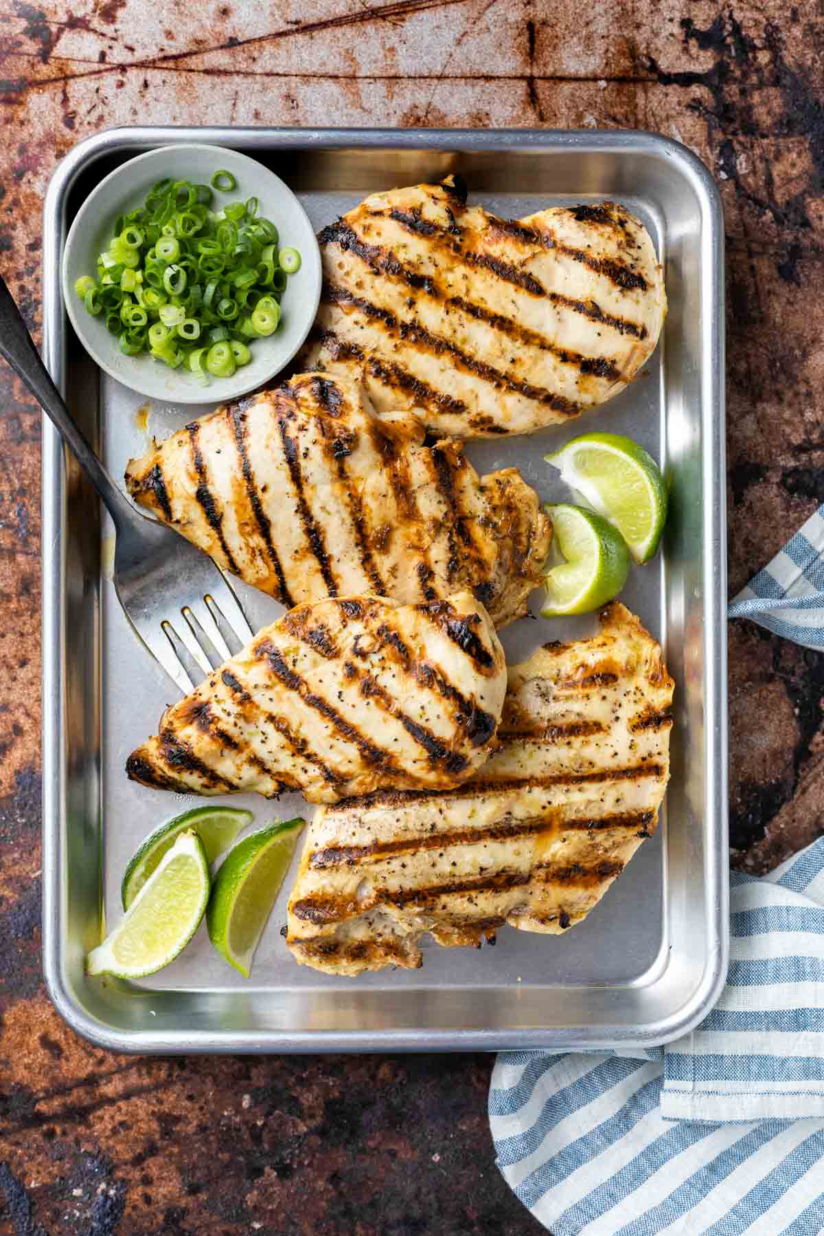 4 grilled chicken breast in a cookie sheet with lime slices and a bowl of green onions 