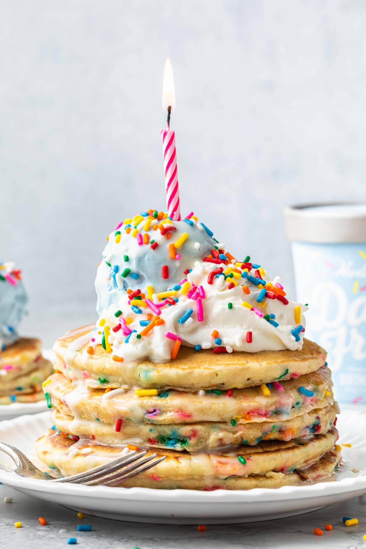 A stack of funfetti pancakes on a plate; topped with whipped cream, blue birthday cake ice cream, rainbow sprinkles, with a birthday candle on top.