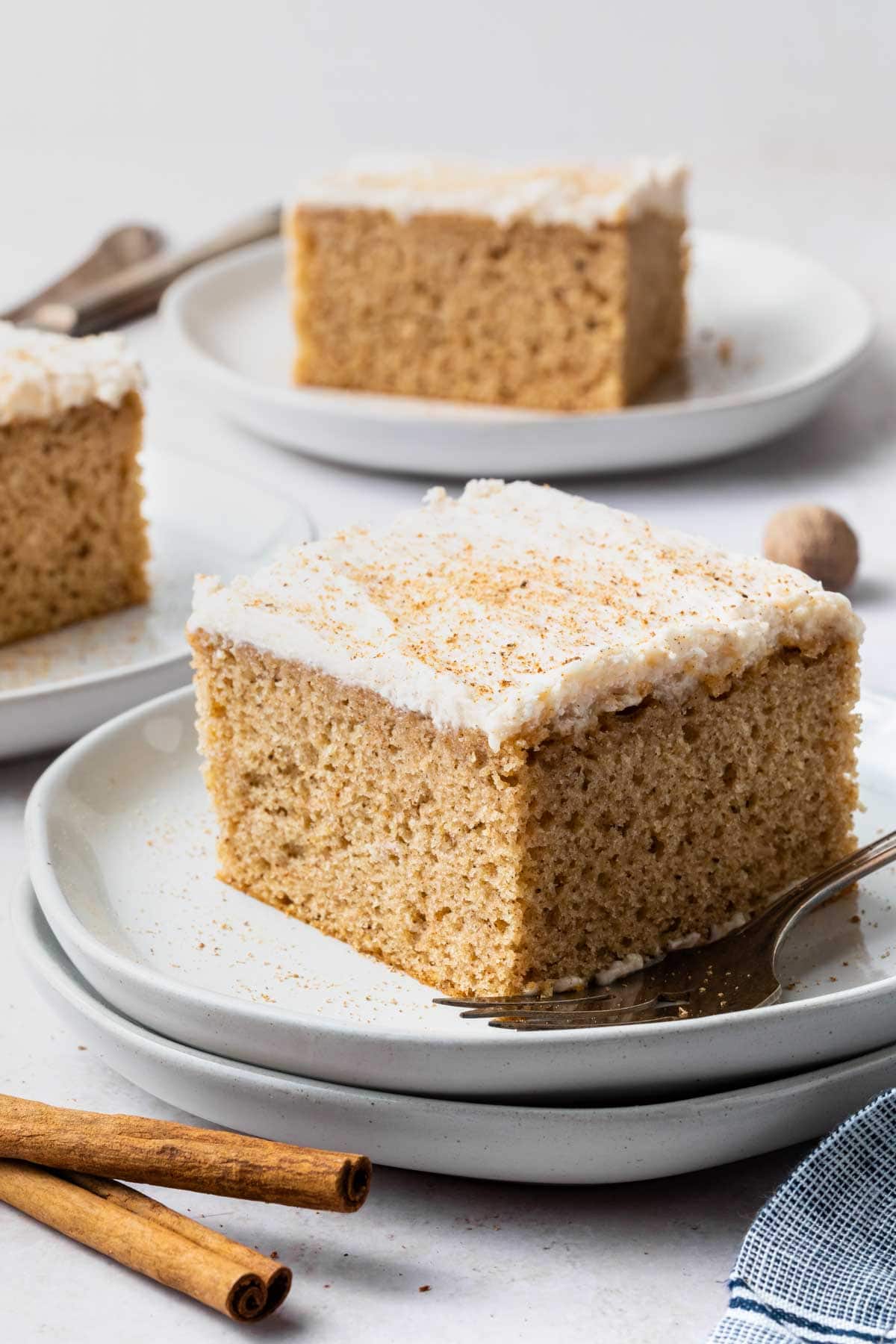 A square of chai spice cake on a white plate with a fork and cinnamon sticks on the counter next to them. 