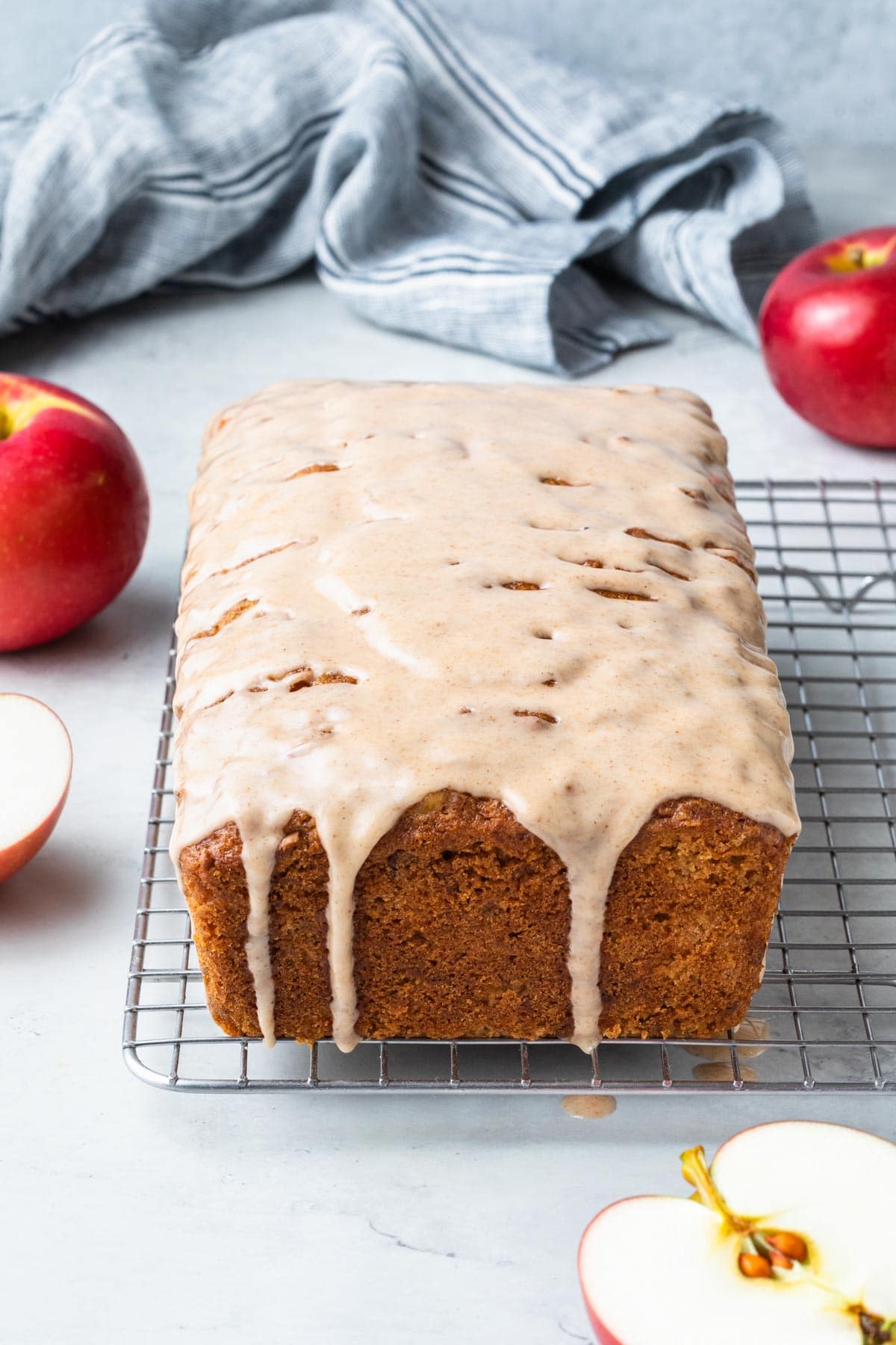 A loaf of apple cinnamon bread covered in a cinnamon glaze with apples around in the background.