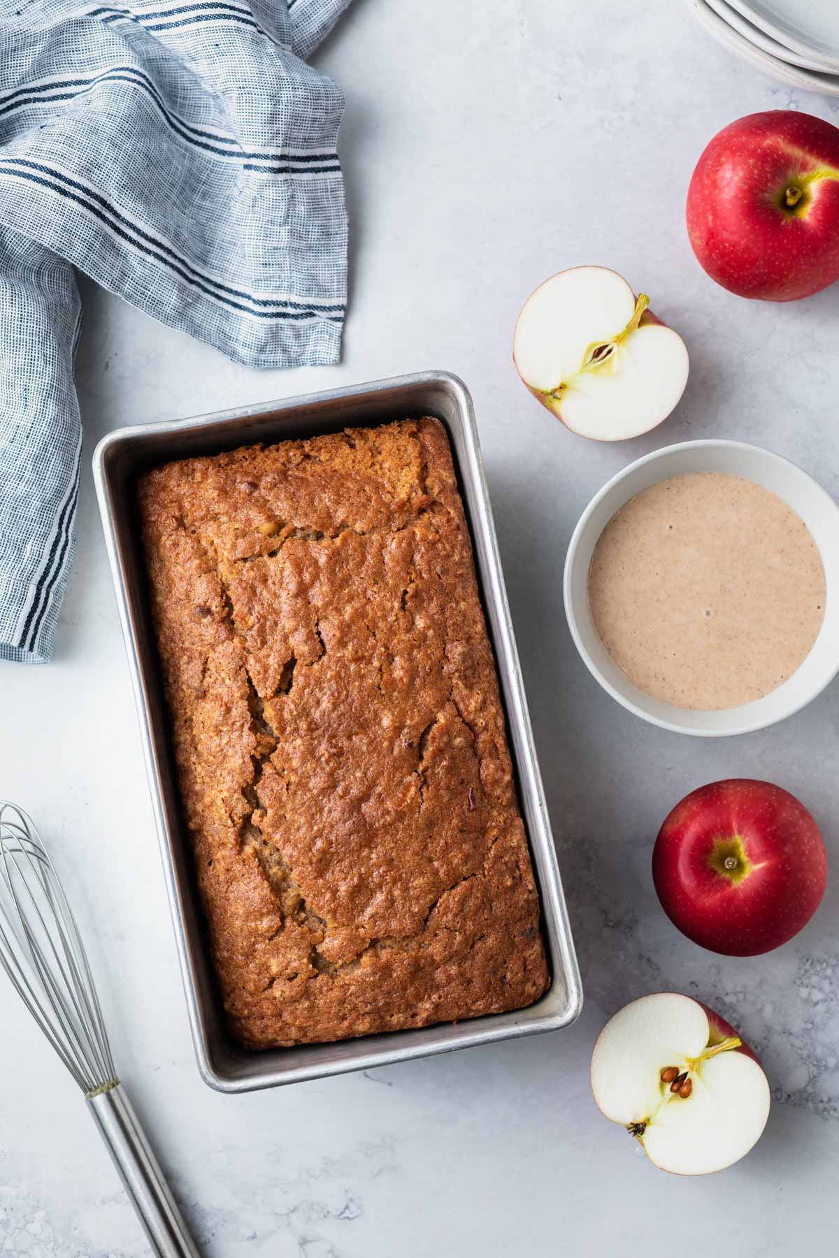 A loaf of apple bread in a metal loaf pan, with a bowl of cinnamon glaze next to it and apples and a whisk to the sides. 