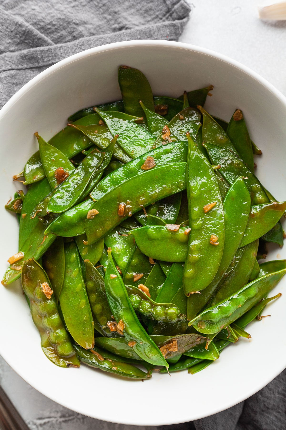 A close up and overhead shot of a bowl of sweet garlicky glazed snow peas with a spoon and some garlic cloves next to the bowl. 