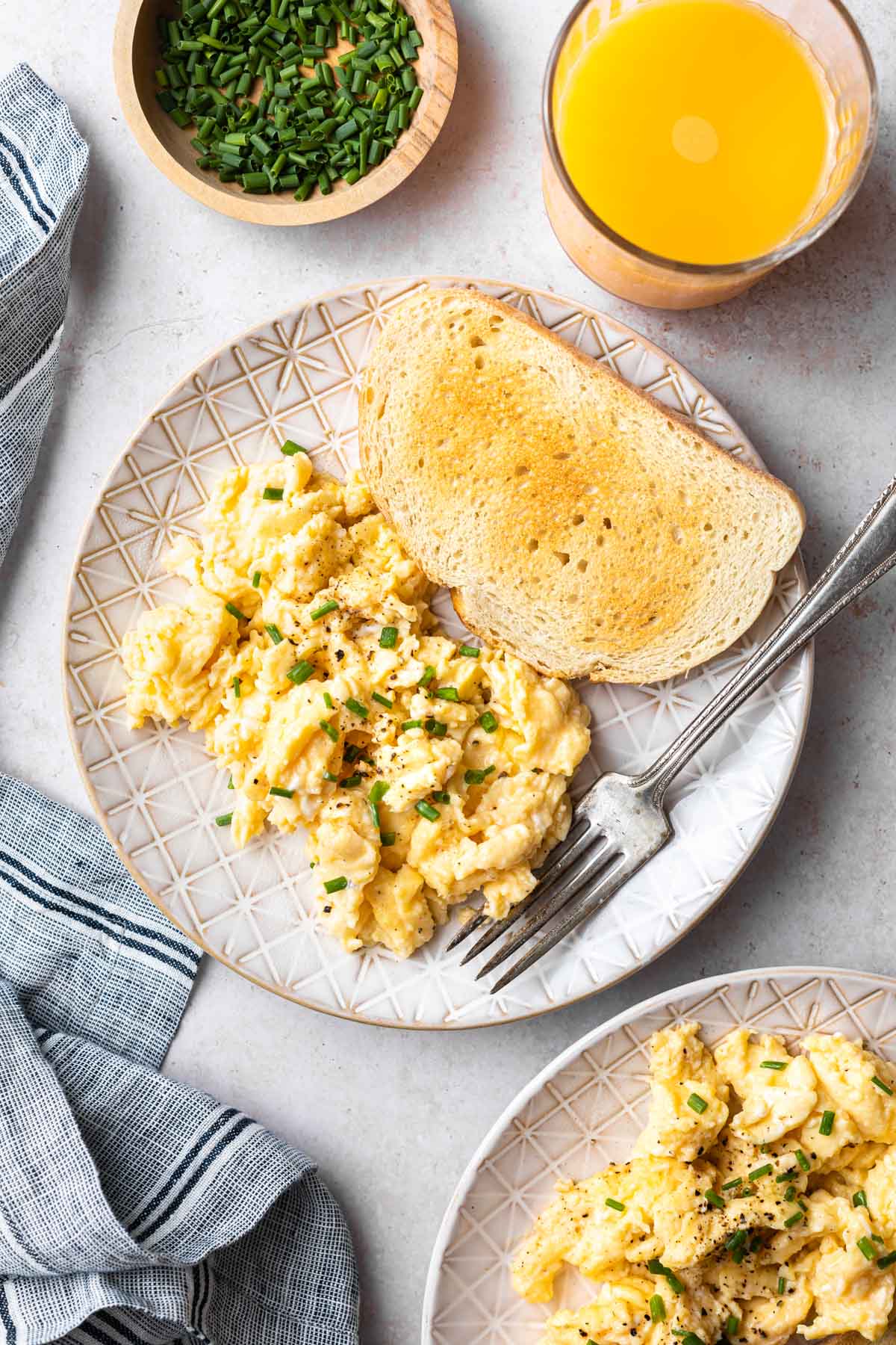 A plate of scrambled eggs sprinkled with chives and toast with a fork next to a glass of orange juice and a small bowl of chopped chives. 