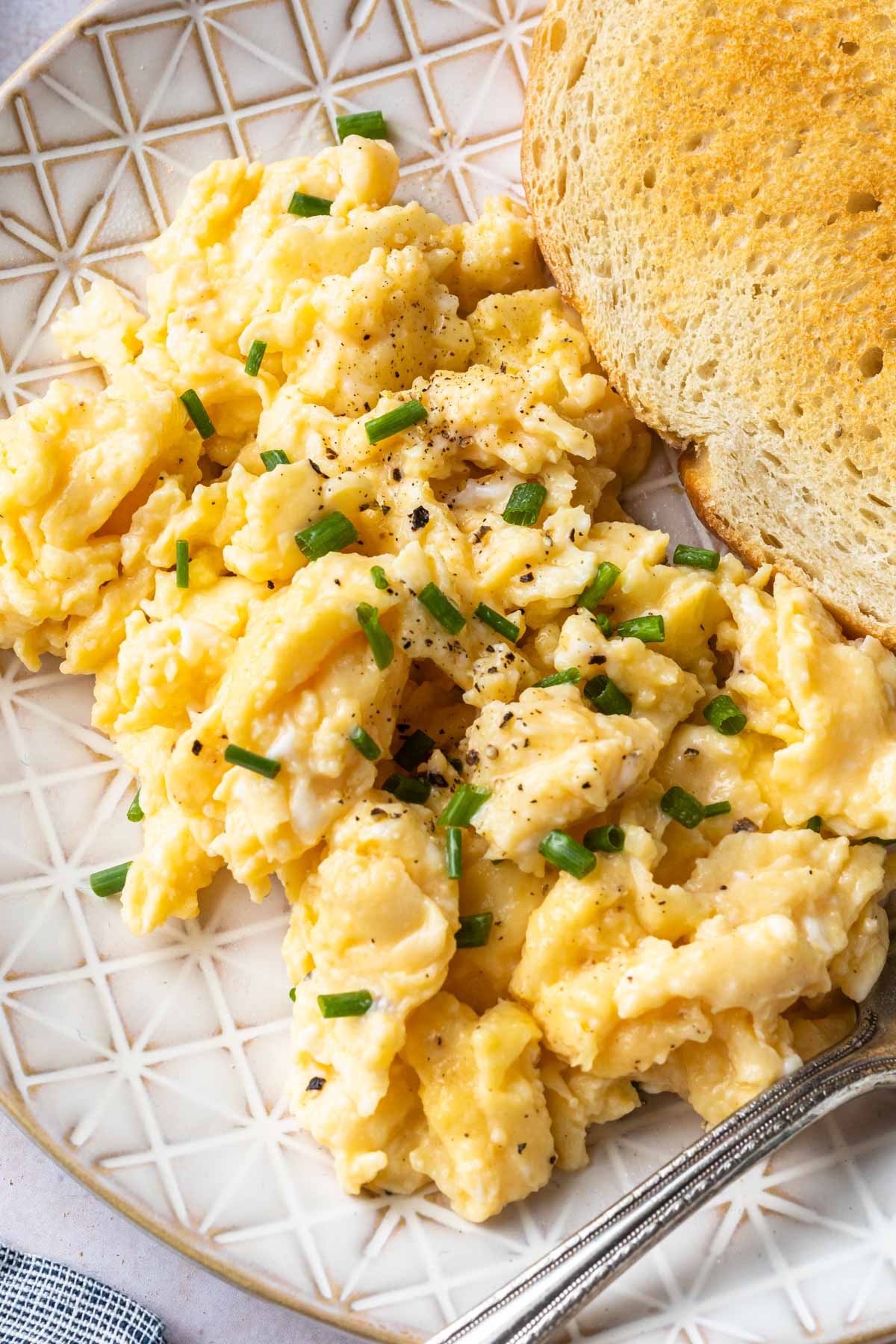 Close up of scrambled eggs sprinkled with salt and pepper and chives on a plate with a fork. 