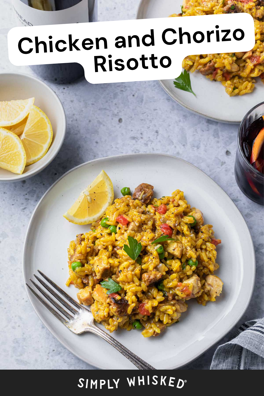 A plate of Chicken and Chorizo Risotto garnished with parsley and a lemon wedge, with a fork beside it. In the background, a glass of drink, lemon slices, and another plate complement this vibrant Chicken and Chorizo Risotto dish.