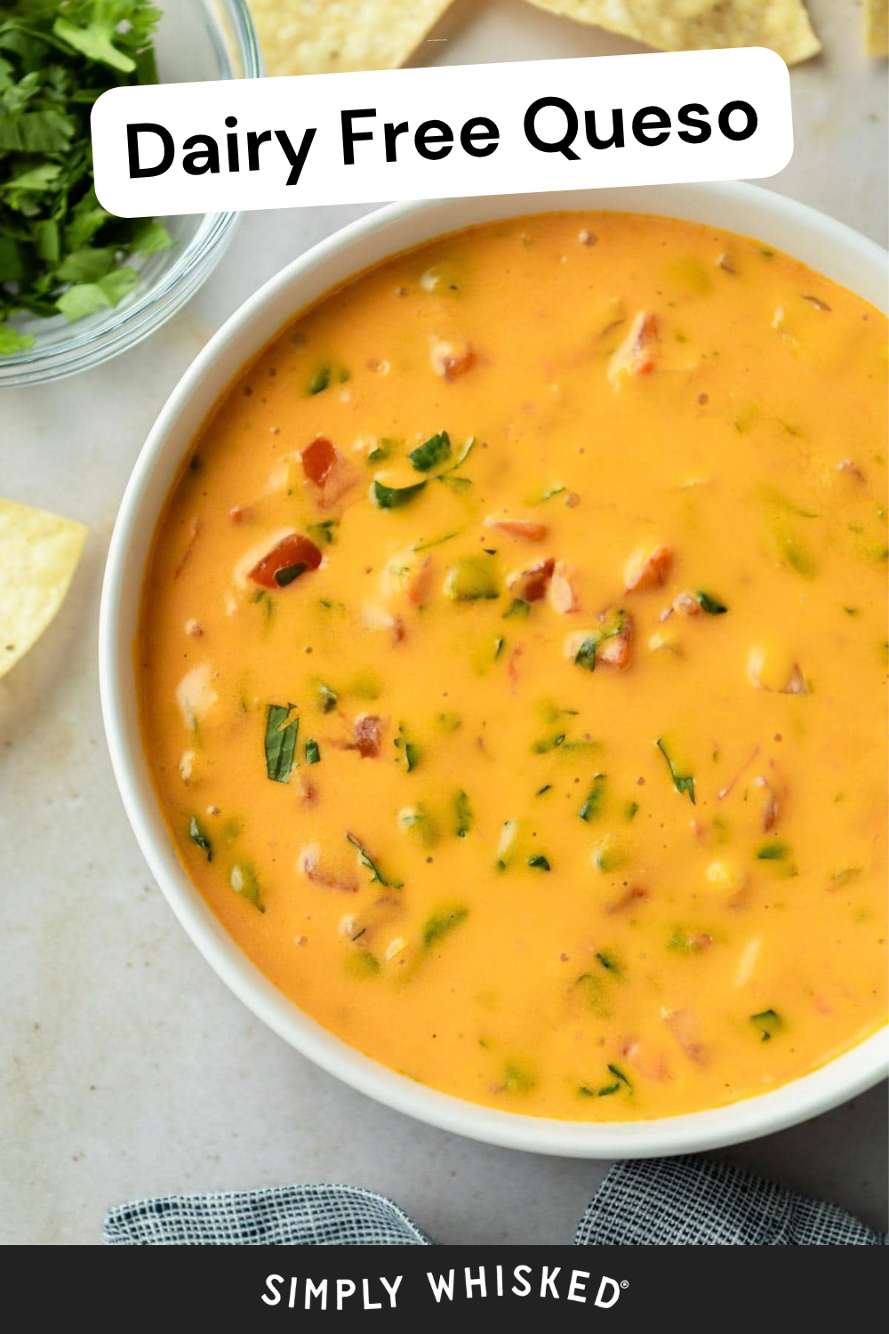 A bowl of creamy orange vegan queso, topped with chopped tomatoes and herbs, is surrounded by crispy tortilla chips and a bowl of fresh cilantro.