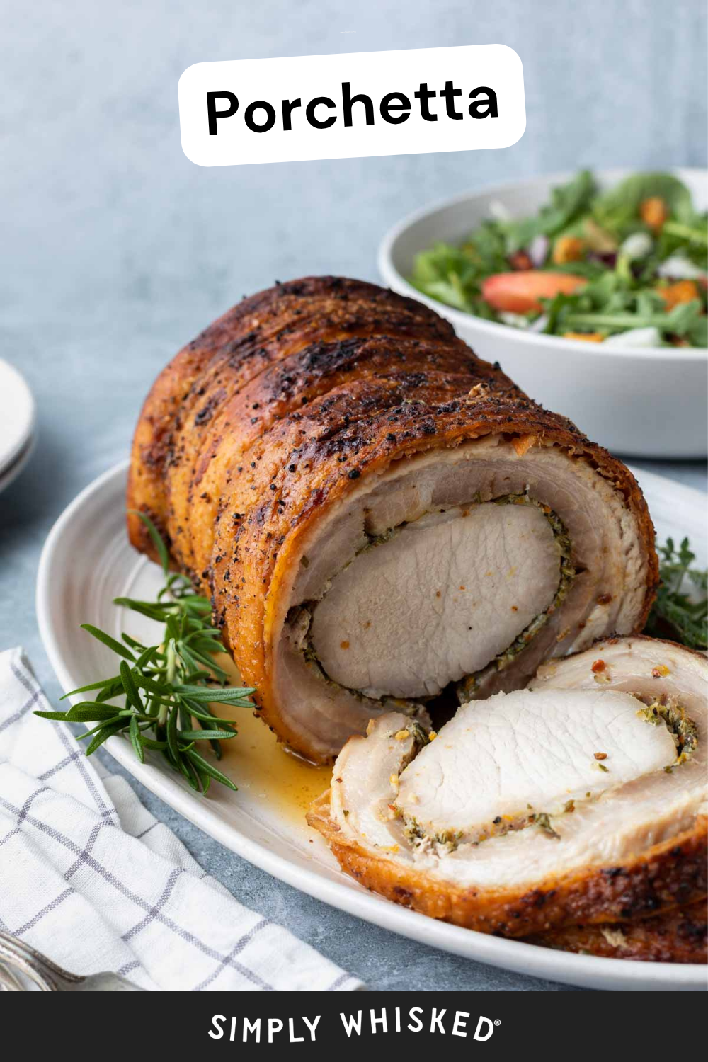 A beautifully sliced porchetta roast on a white platter, garnished with fresh rosemary, with a vibrant salad in the background and a striped napkin nearby.