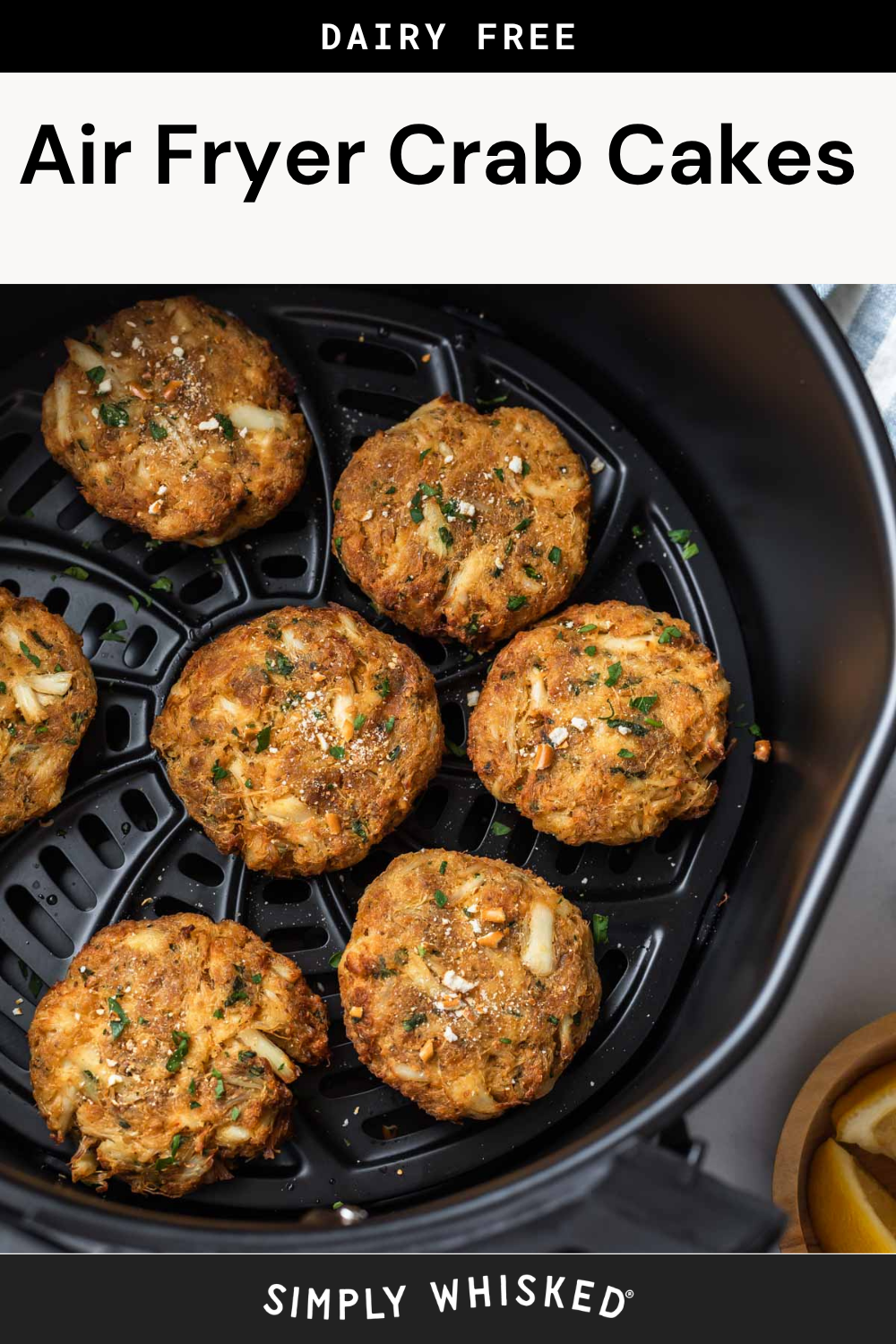 Six golden-brown air fryer crab cakes sit in an air fryer basket, garnished with fresh herbs and accompanied by lemon wedges in the corner. The image features text at the top that reads “Dairy Free Air Fryer Crab Cakes.”.